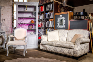 A sitting room that highlights one textured, white couch, a white chair, and a white bookshelf with a pink light.